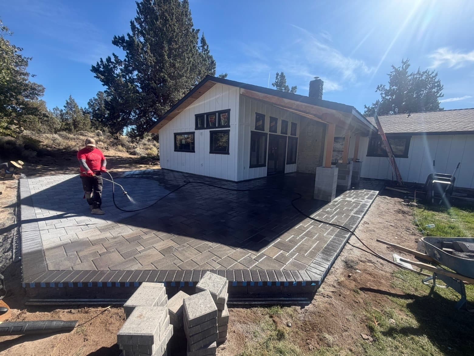 Back patio with rectangular pavers, a dark border, and masonry column bases beside a covered porch; a worker rinses the surface.