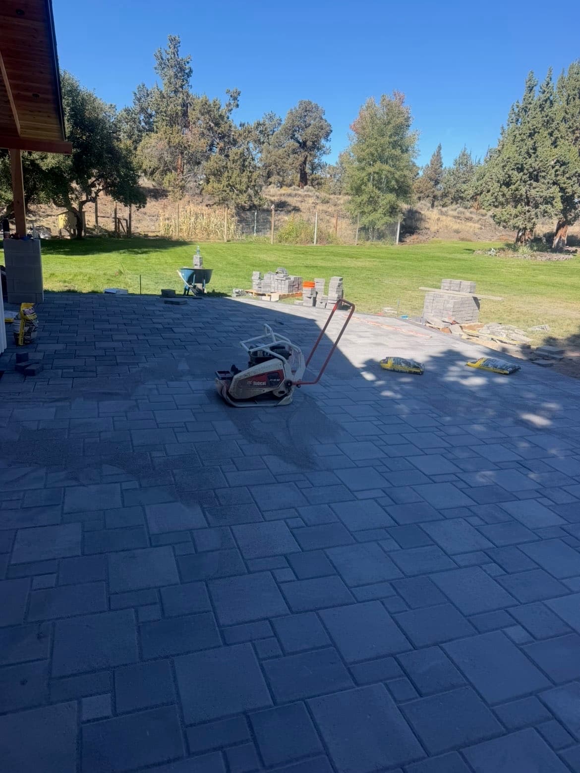 View from the covered porch across the rectangular patio pavers toward the lawn, with a plate compactor and stacked materials on site.