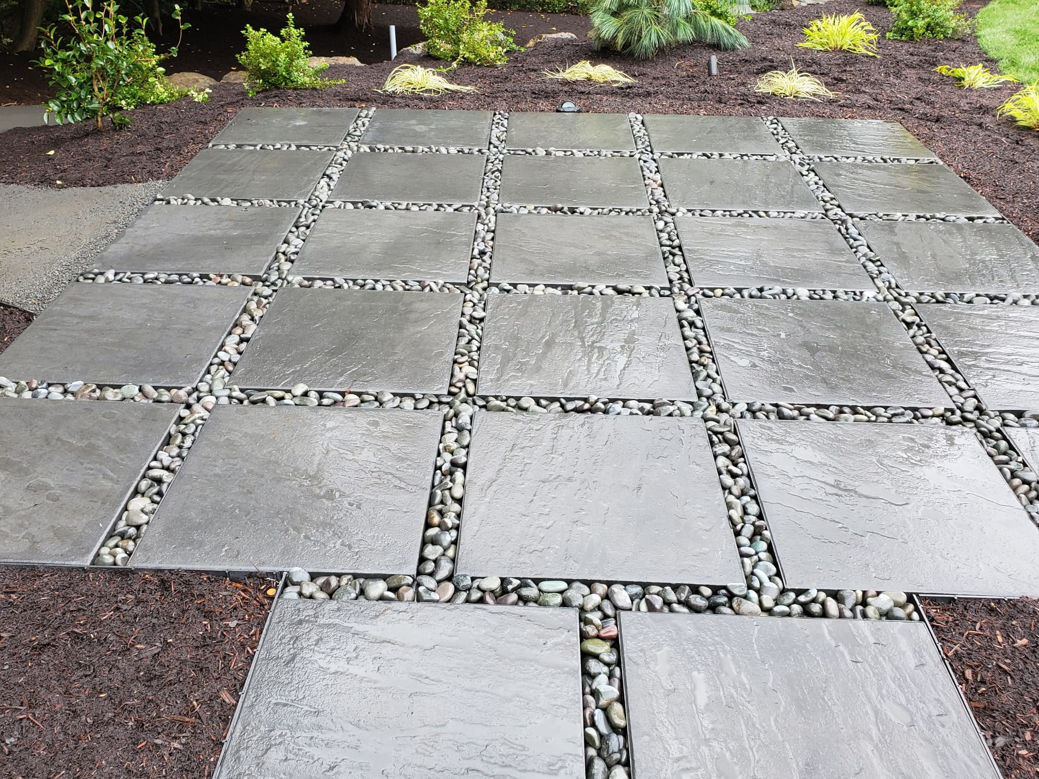 Large square pavers set in decorative river rock beside planted garden beds.